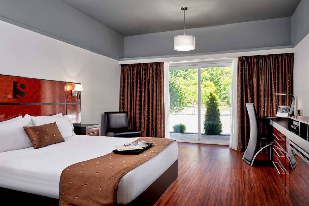 A modern hotel room with a large bed, a desk, and a view of greenery outside.