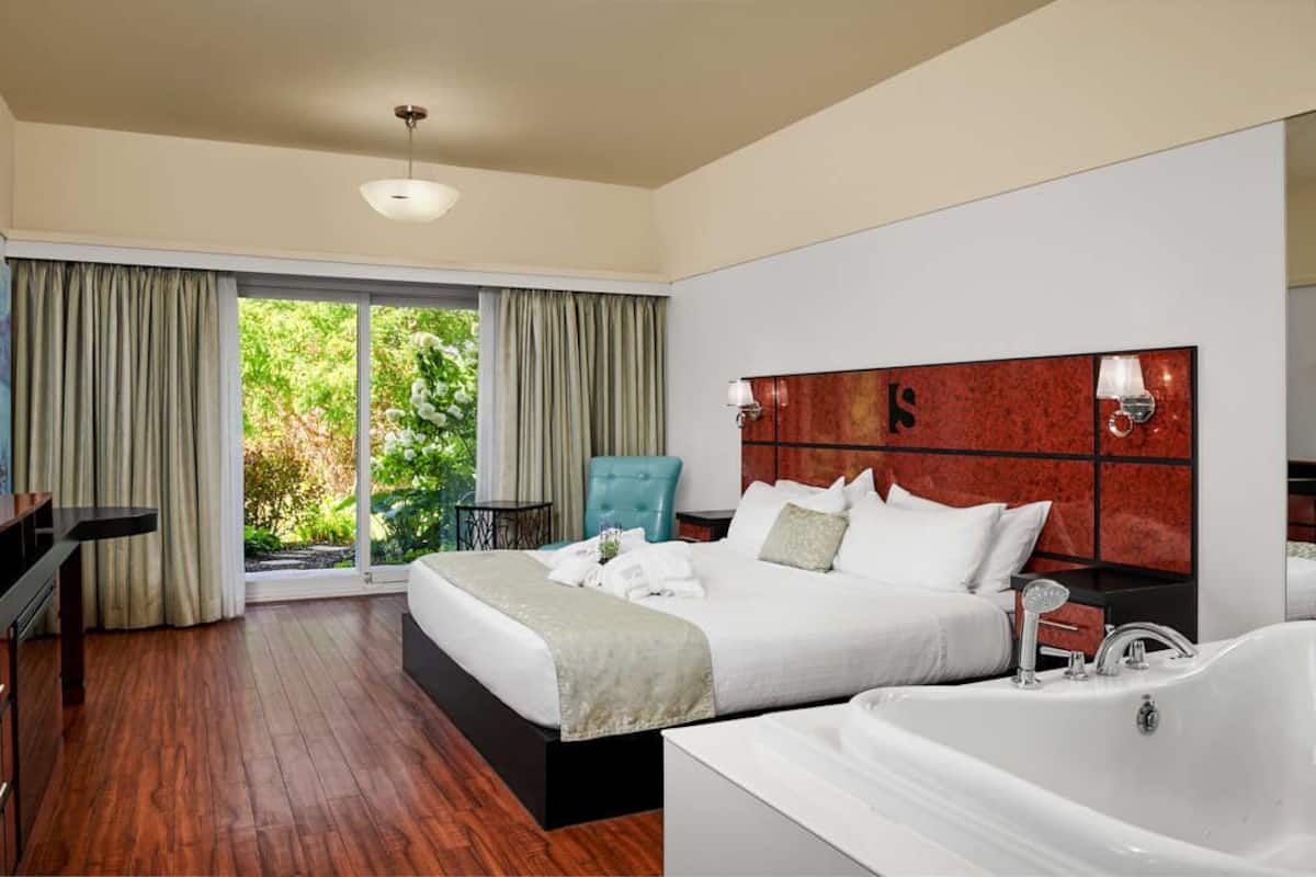 A modern hotel room with a large bed, a separate sitting area with a chair, a bathtub, and a view of greenery through the windows.