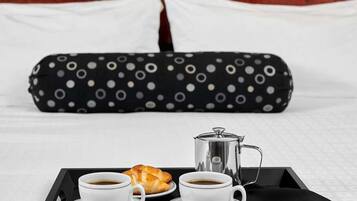Un plateau de petit-déjeuner bien présenté, avec deux tasses de café, un croissant et une cafetière, le tout posé sur un plateau noir, sur un lit agrémenté d’oreillers blancs et d’une taie d’oreiller à pois.