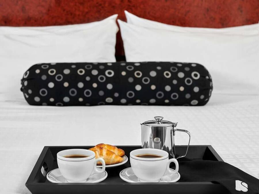Un plateau de petit-déjeuner bien présenté, avec deux tasses de café, un croissant et une cafetière, le tout posé sur un plateau noir, sur un lit agrémenté d’oreillers blancs et d’une taie d’oreiller à pois.