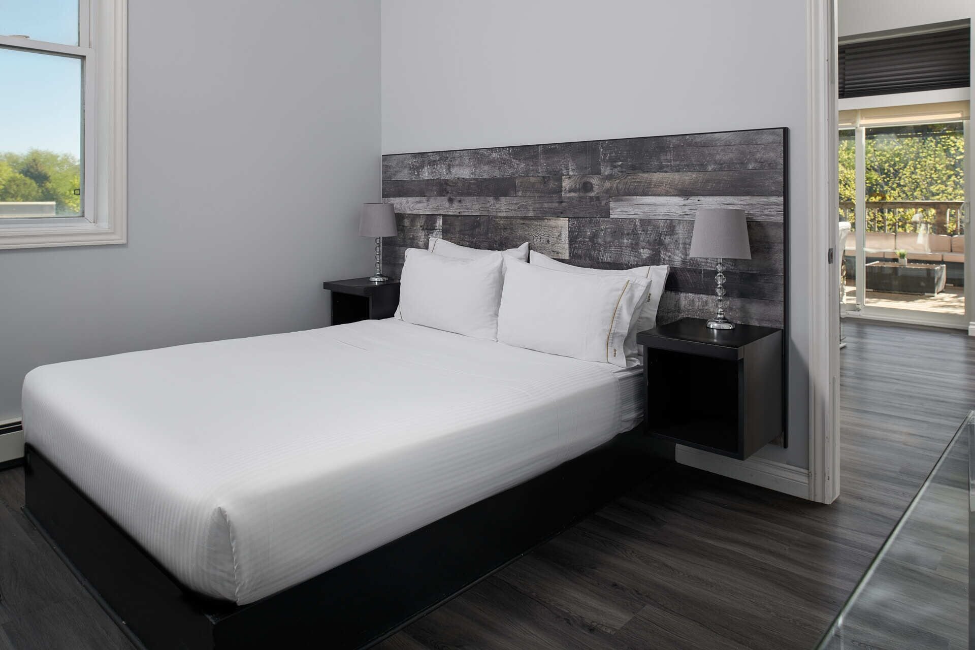 Quarto moderno com uma cama grande, piso de madeira escura e vista para o exterior por uma porta de vidro.