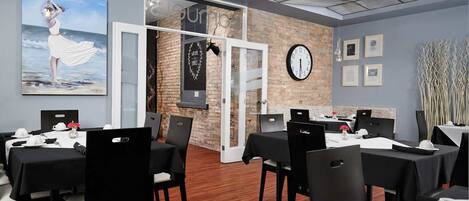 Un espace de restauration moderne, avec des tables dressées pour le service, une horloge au mur et une vue sur la plage par une porte ouverte.