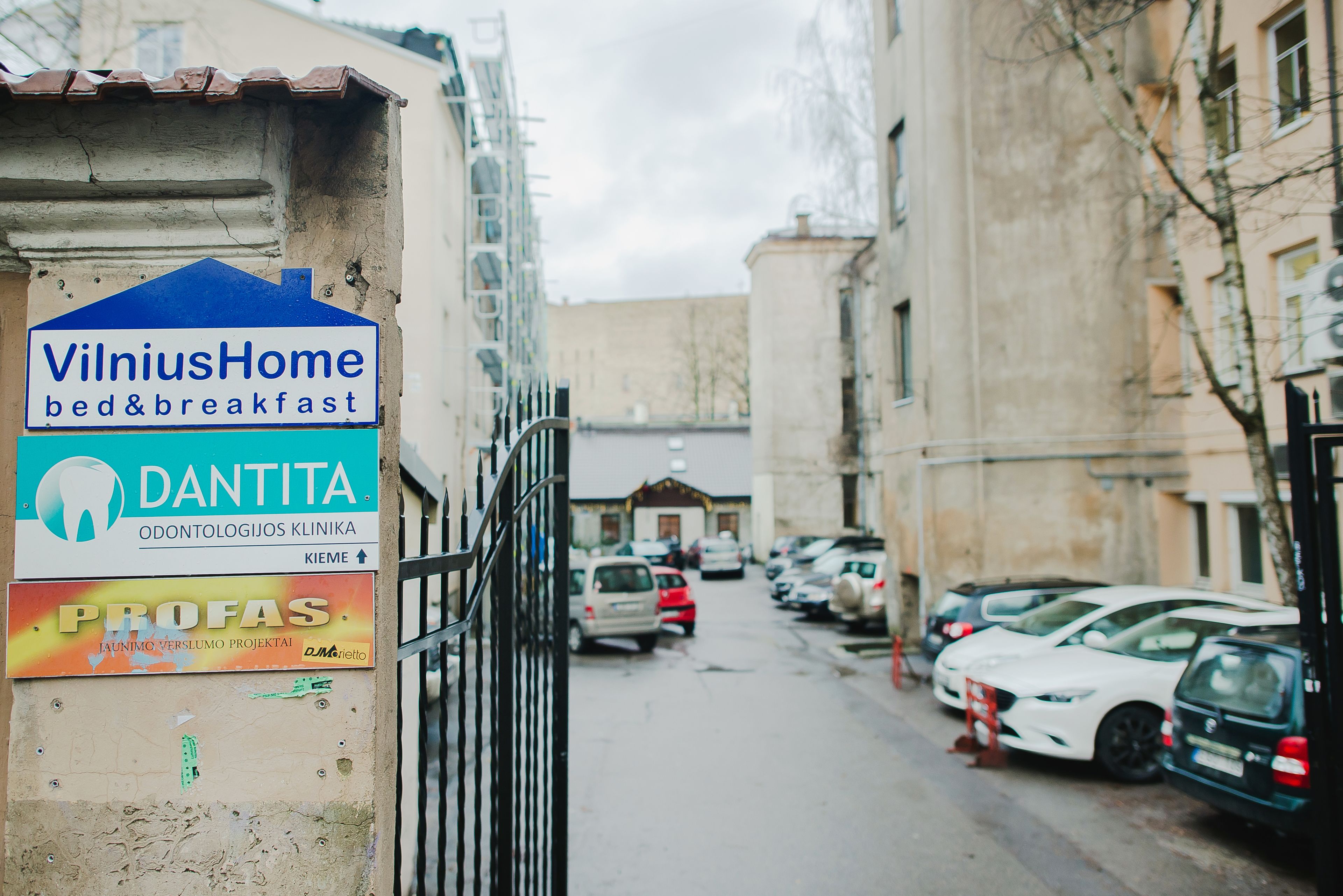 A narrow street with parked cars and buildings on either side. A sign for 'VilniusHome bed&breakfast' and 'Dantita Odontologijos Klinika' is visible.