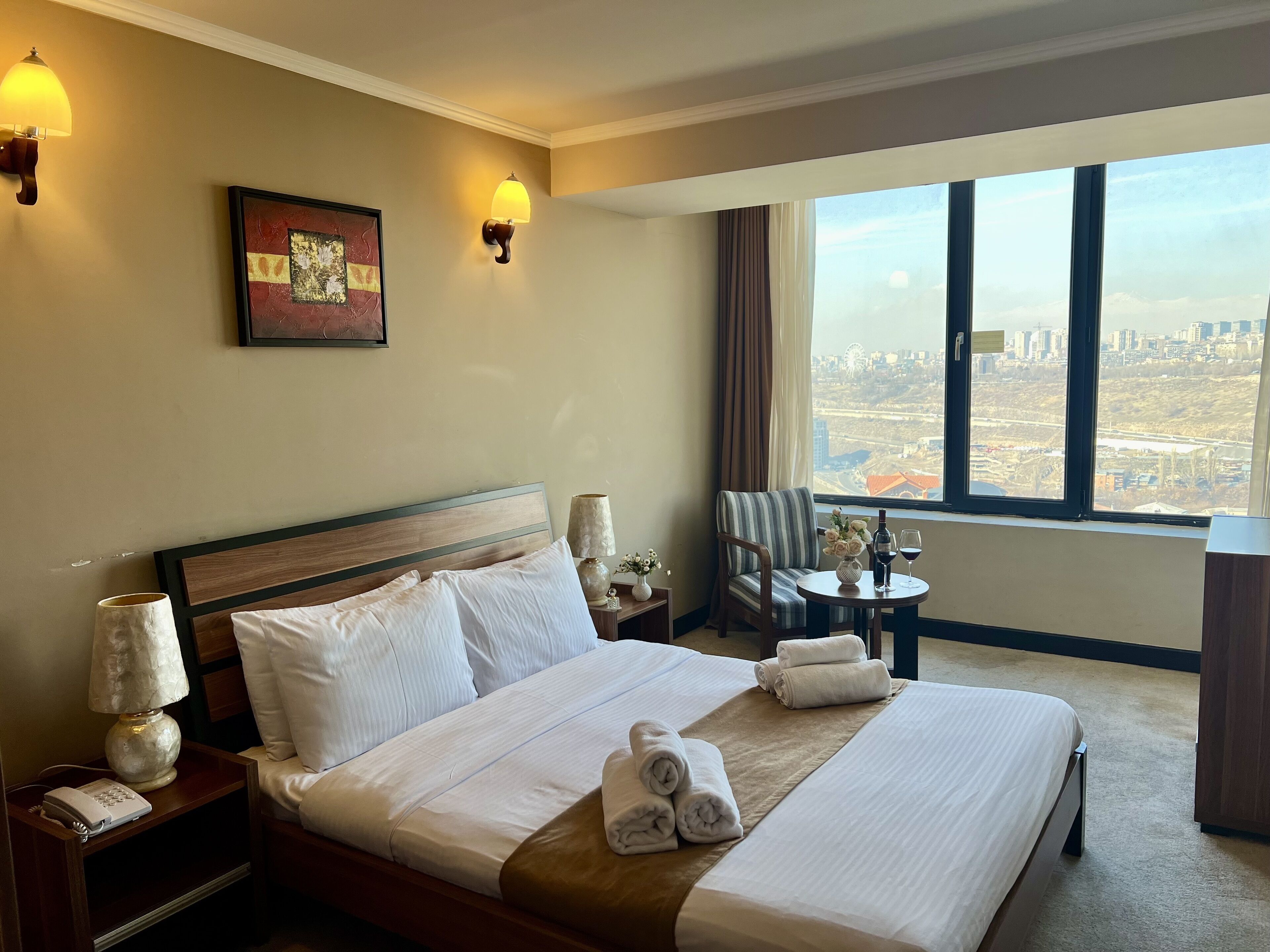 Habitación de hotel con una cama grande, una silla, una mesita con un florero y copas de vino, y vistas de la ciudad a través de amplios ventanales.