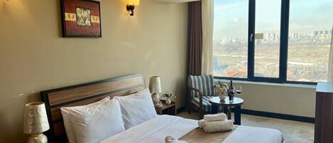 Habitación de hotel con una cama grande, una silla, una mesita con un florero y copas de vino, y vistas de la ciudad a través de amplios ventanales.