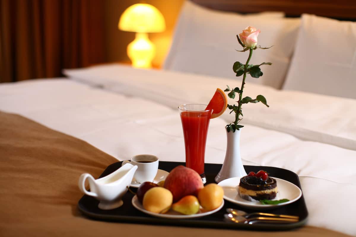 Un plateau contenant des aliments pour le petit-déjeuner, notamment des fruits, un verre de jus, une tasse de café et un dessert, posé sur le lit d’un hôtel.