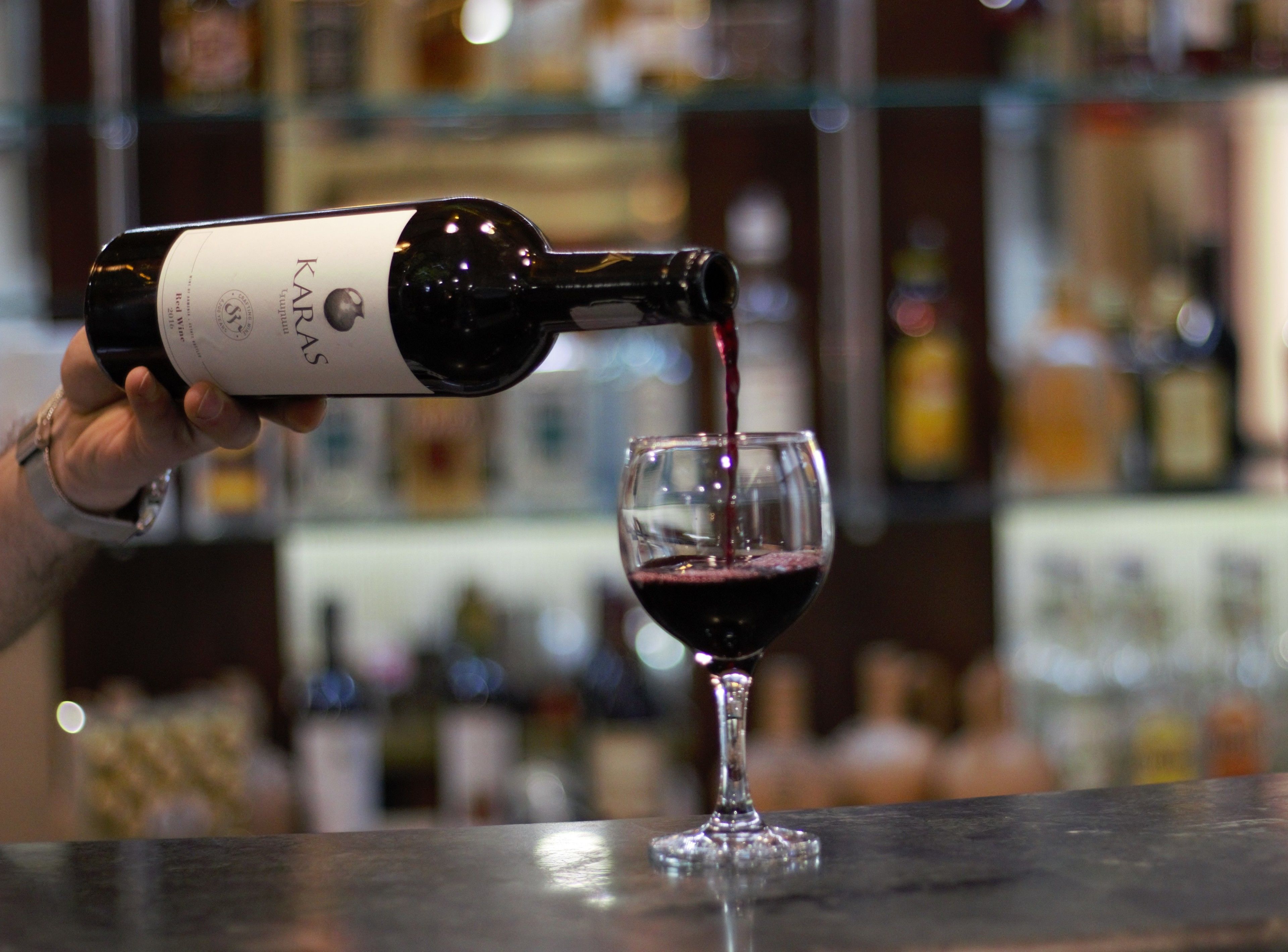 Una persona sirve vino tinto de una botella con la etiqueta 'KARAS' en una copa.