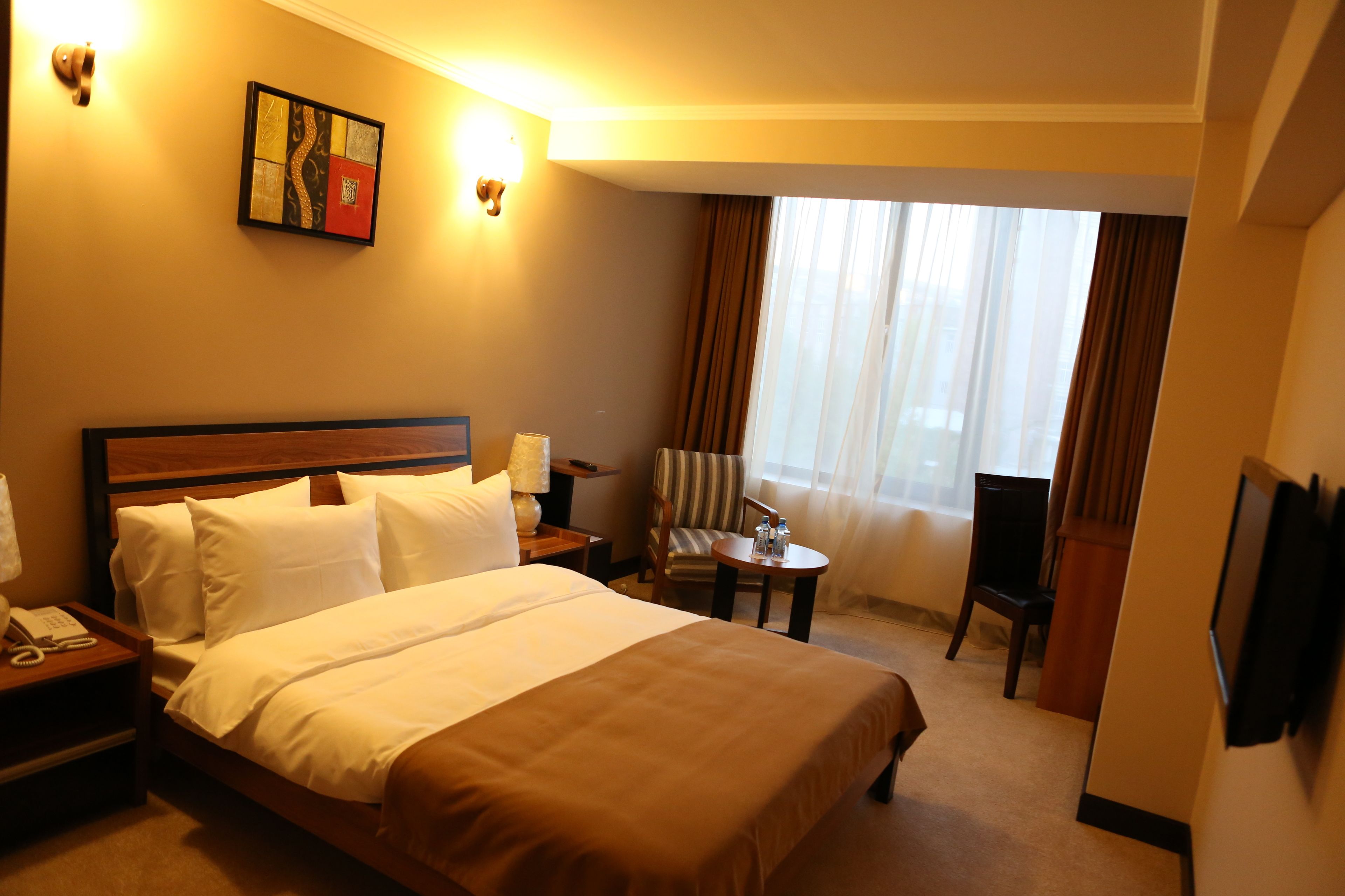 Habitación de hotel con una cama grande, una mesita de noche con lámpara, una silla, una pequeña mesa con una botella y un televisor montado en la pared.
