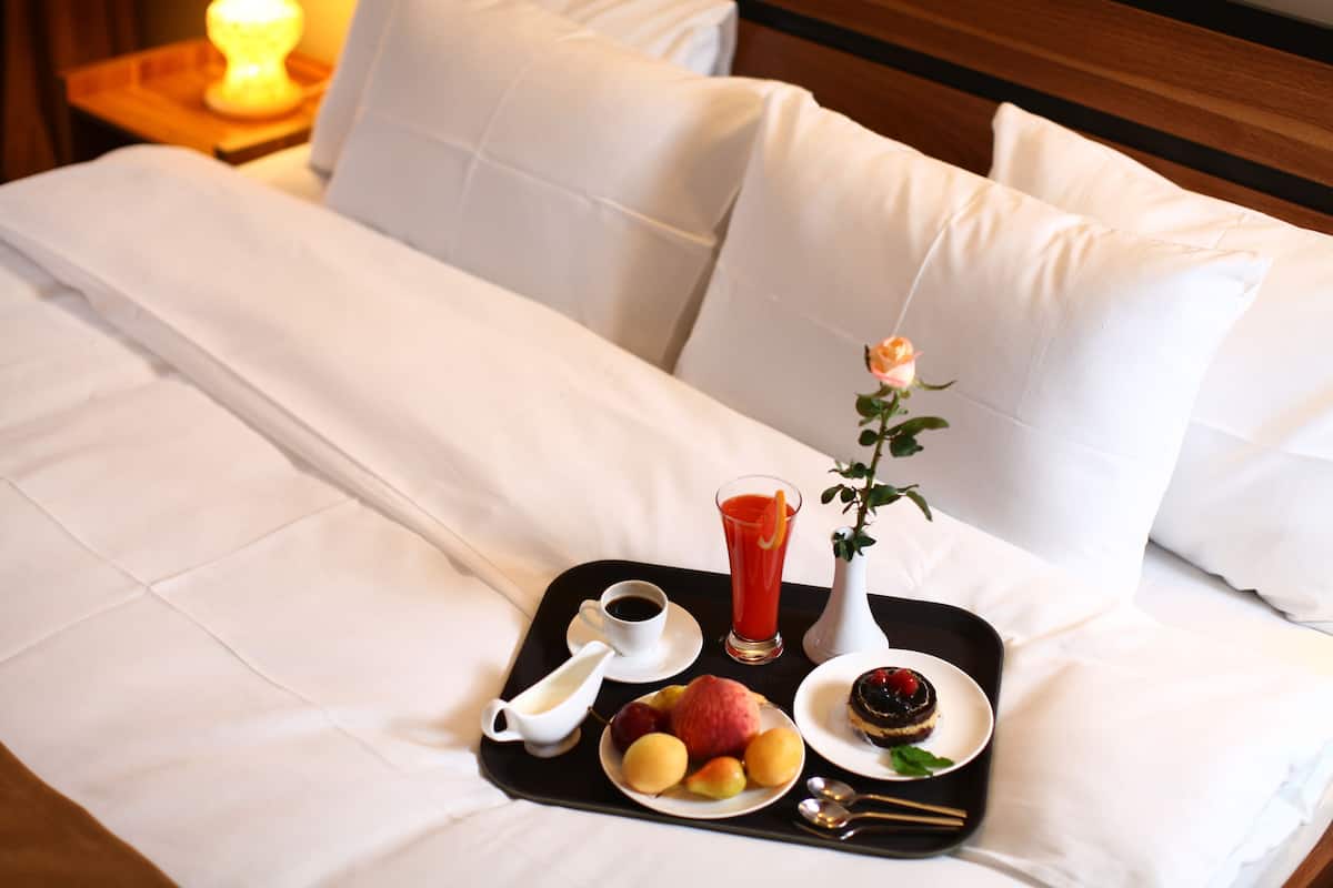 Un lit bien fait, avec du linge de lit blanc, un plateau contenant des aliments pour le petit-déjeuner, notamment des fruits, un verre de jus et un petit vase avec une fleur.