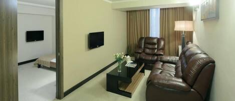 A hotel room with a brown leather sofa, a reclining chair, a flat-screen TV mounted on the wall, a small table with a vase, and a bed in the background.