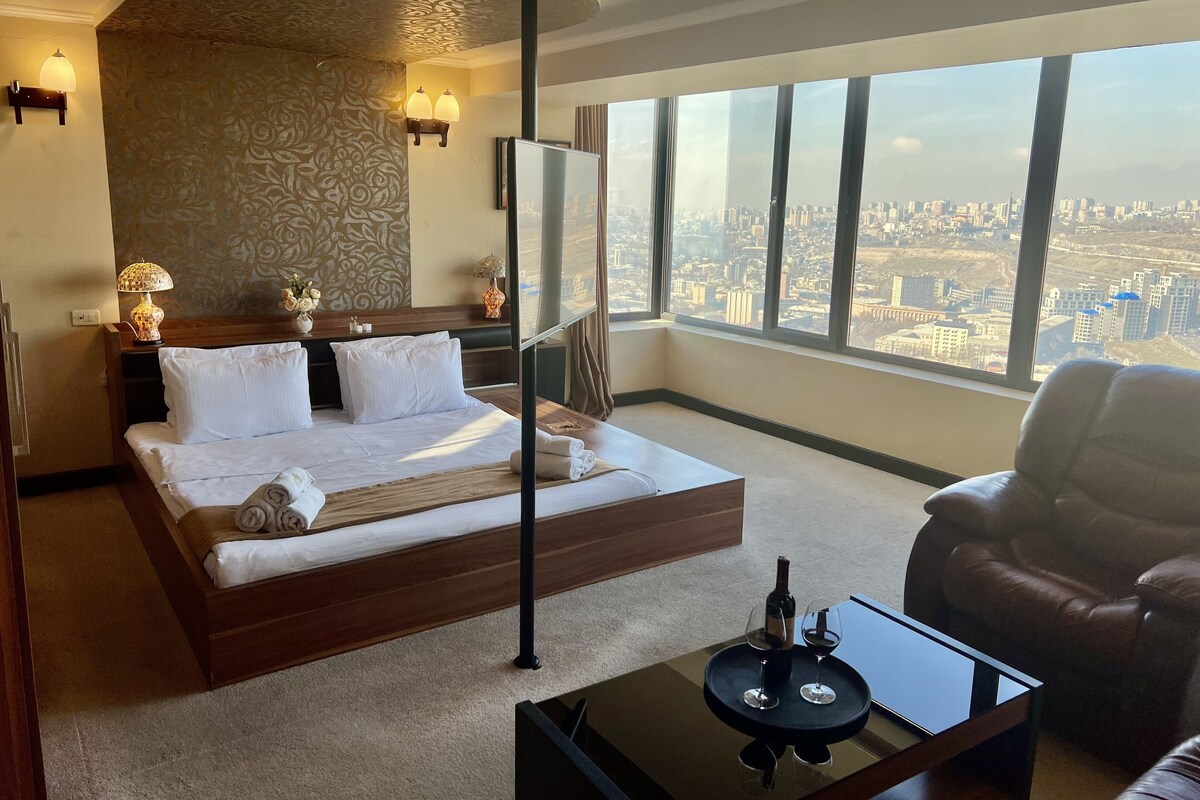A hotel room with a large bed, a view of the city, and a leather armchair.