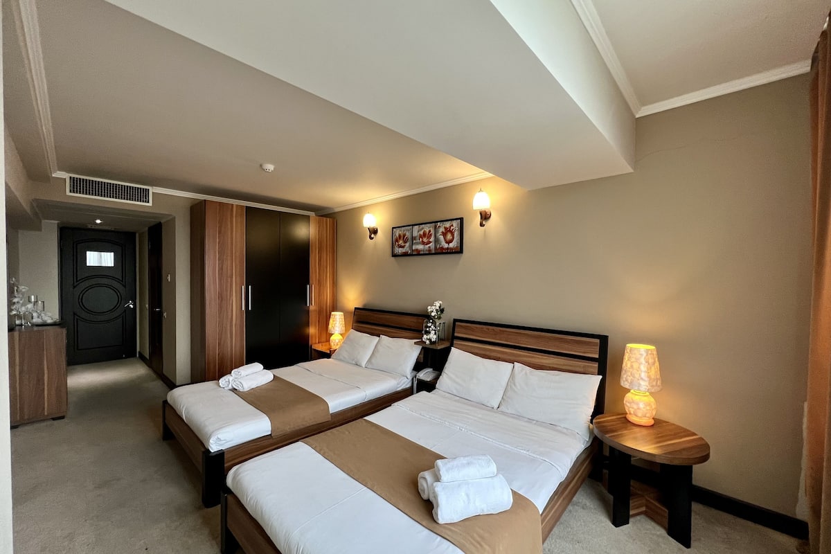 Une chambre d’hôtel avec deux lits, une tête de lit en bois, une table de chevet avec une lampe et une porte dotée d’une grille d’aération.