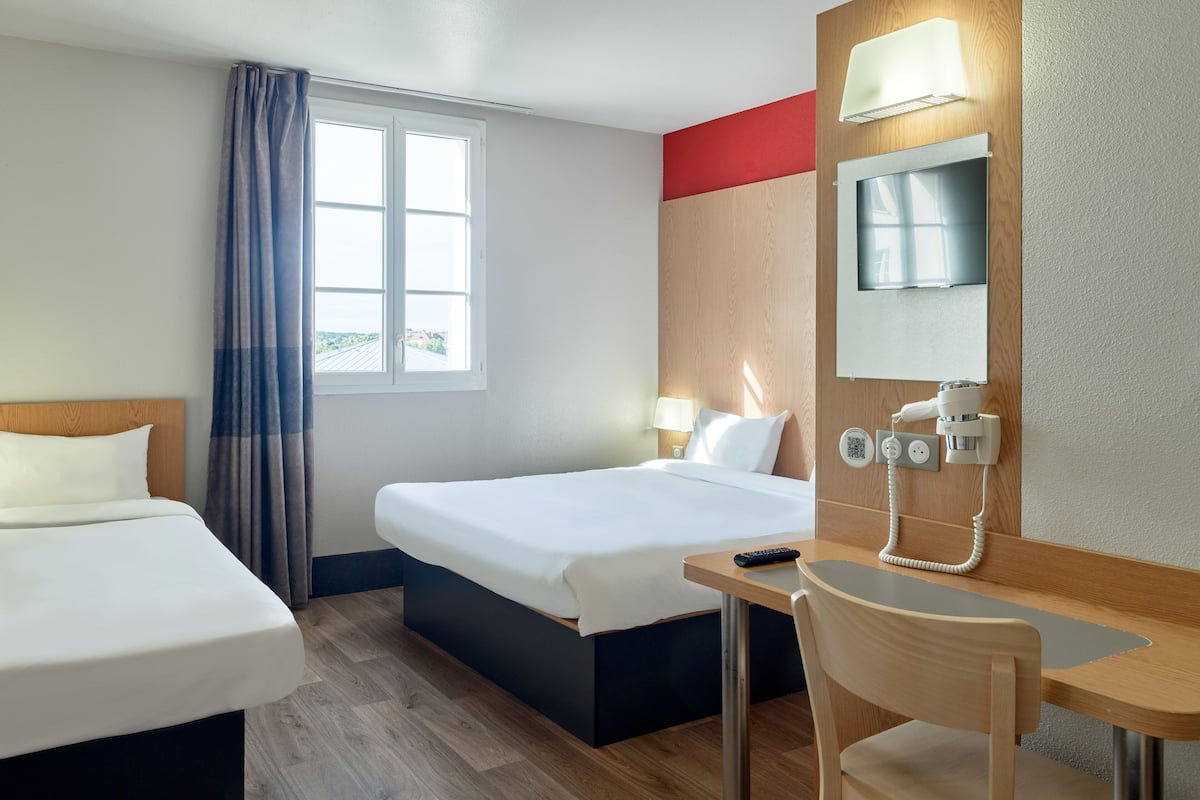 Une chambre d’hôtel avec deux lits, un bureau avec une chaise, une lampe de bureau, un téléphone et une fenêtre avec des rideaux.