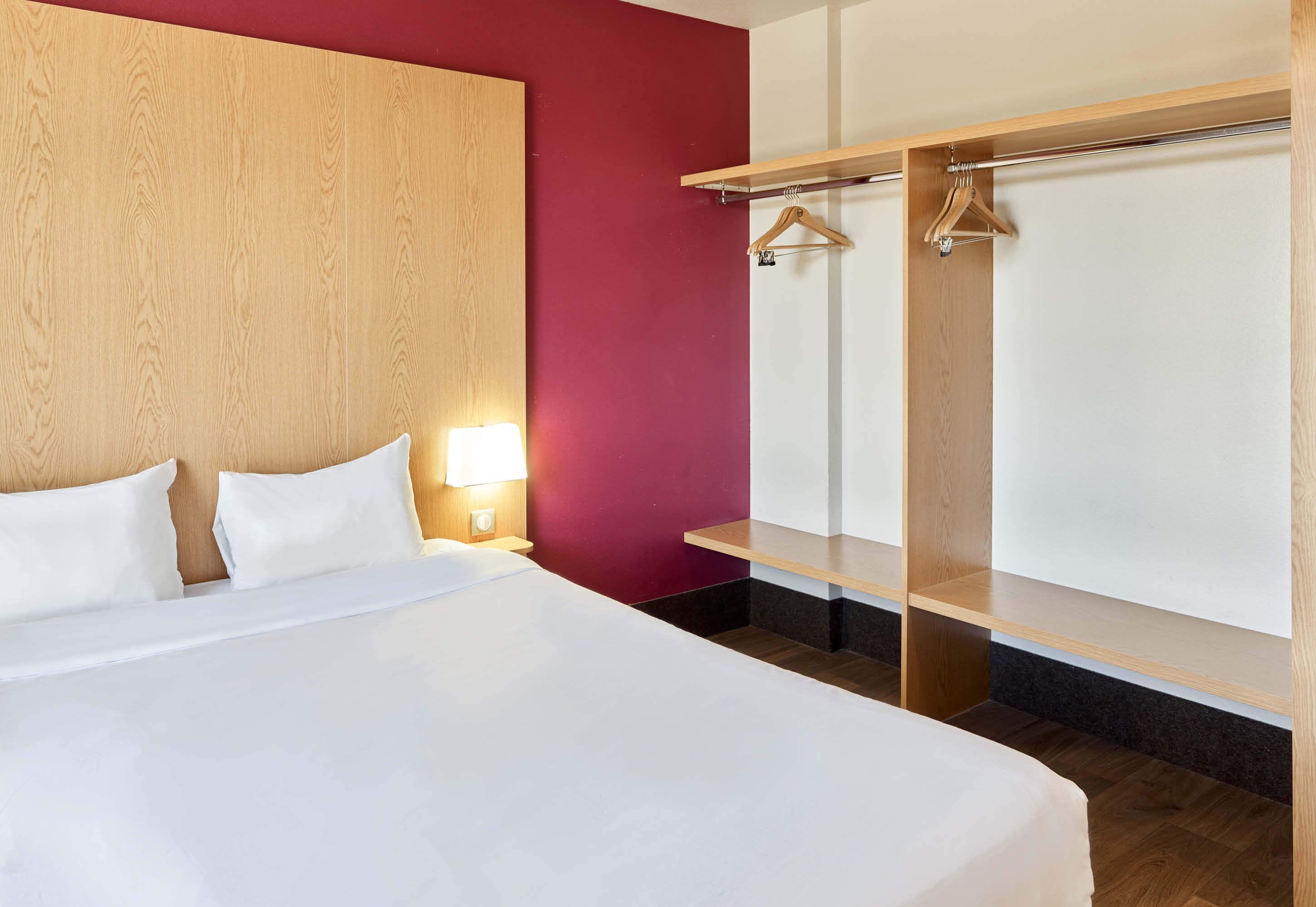 A hotel room with a large bed, wooden headboard, a red accent wall, a wardrobe with a mirror, and a nightstand with a lamp.