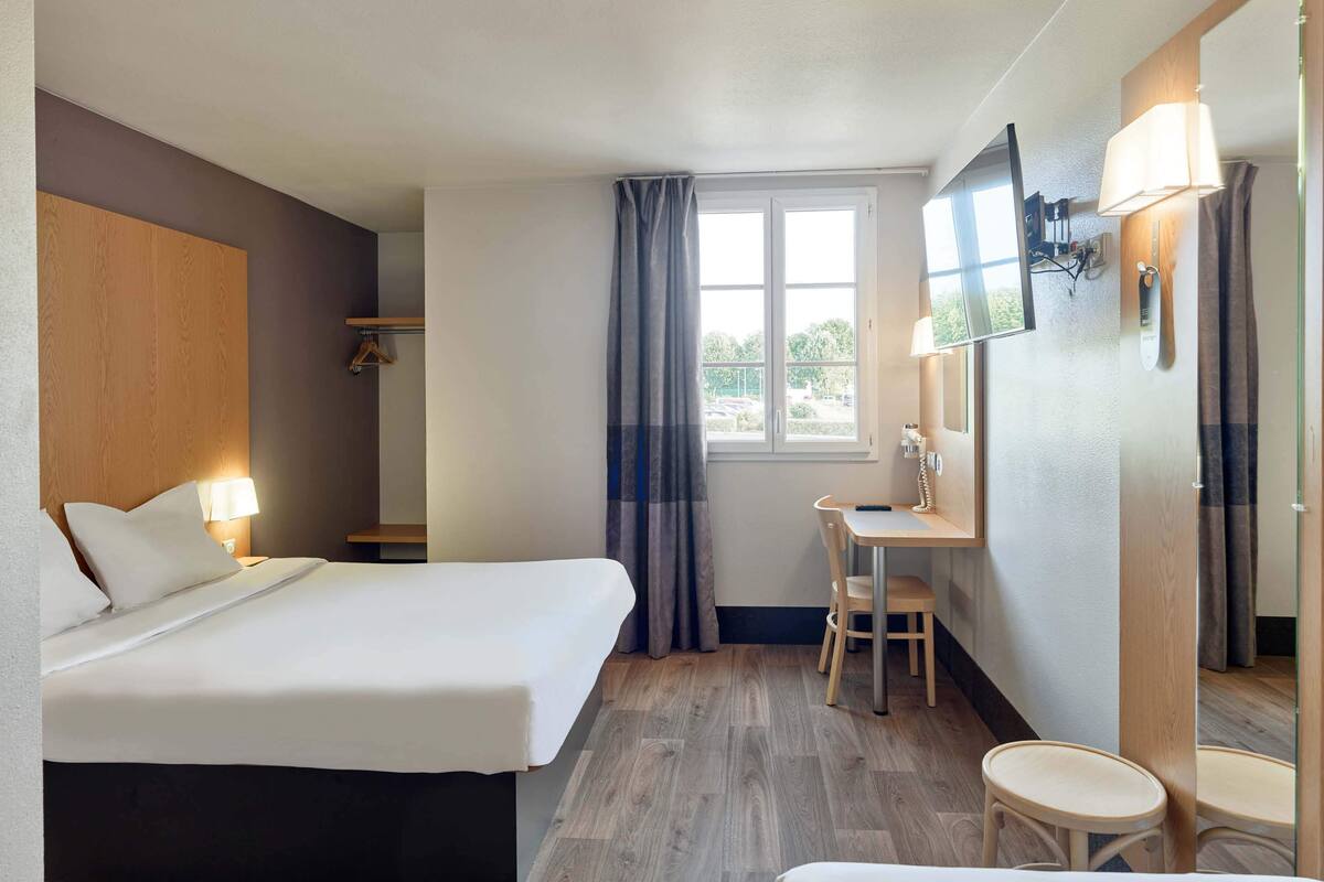 Une chambre d’hôtel avec deux lits, un bureau, une chaise, un petit tabouret, un miroir et une fenêtre avec des rideaux.