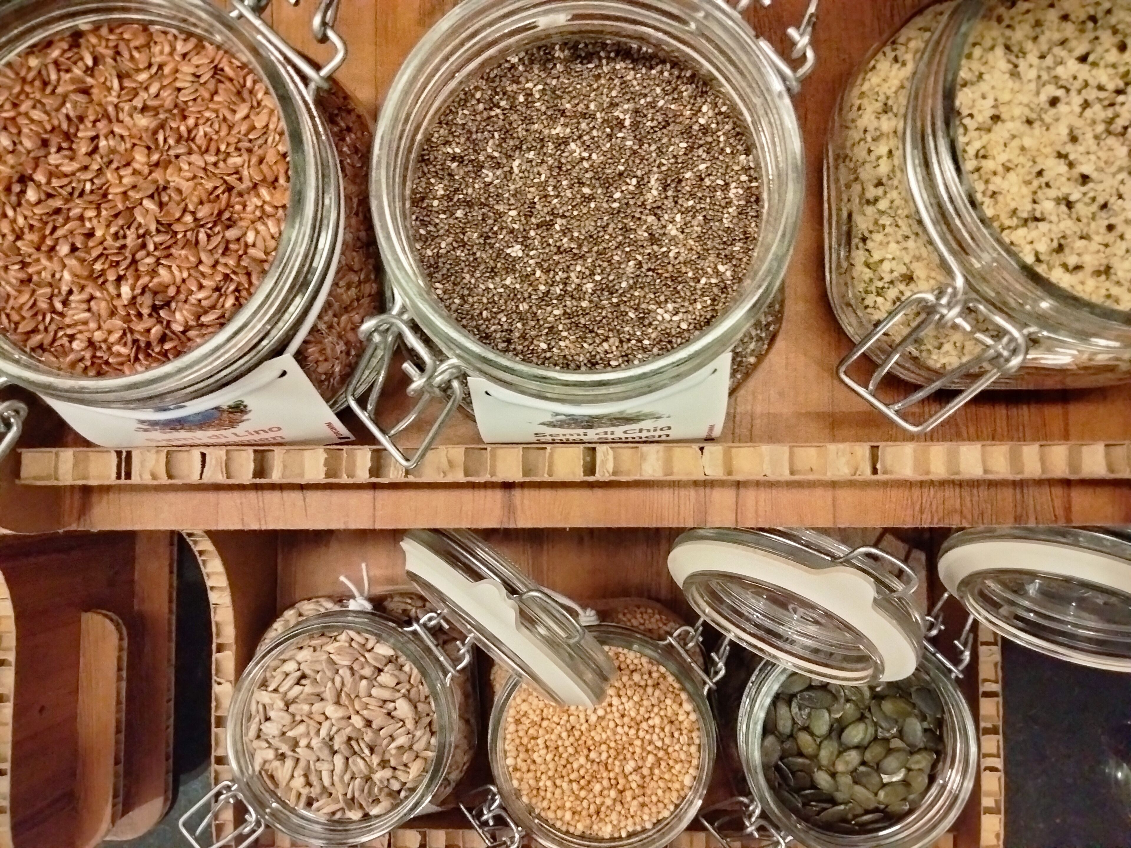 A variety of glass jars containing different types of seeds and grains, organized on wooden shelves.
