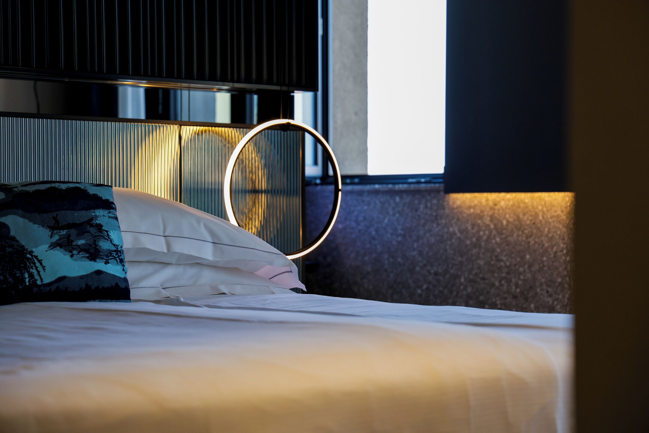 Uma cama com luminária circular acima, travesseiro com desenho de natureza e janela com persianas.