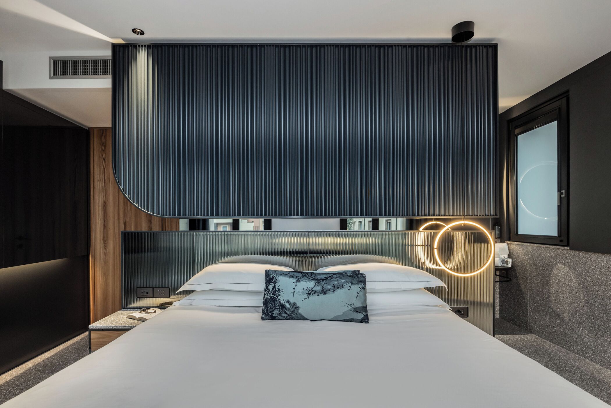 Ein modernes Schlafzimmer mit einem großen Bett, einer Kopfteil mit vertikalen Linien, einem Nachttisch mit Lampe und Blick ins Freie.