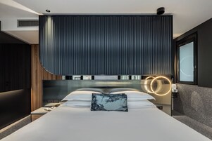 Una camera da letto moderna con un letto grande, una testiera con linee verticali, un comodino con una lampada e vista sull'esterno.