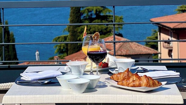 A table set with a croissant, a glass of red wine, a glass of orange juice, and several white cups and saucers. - Hotel Al Caminetto – Adults Only, WorldHotels Crafted (Torri del Benaco)