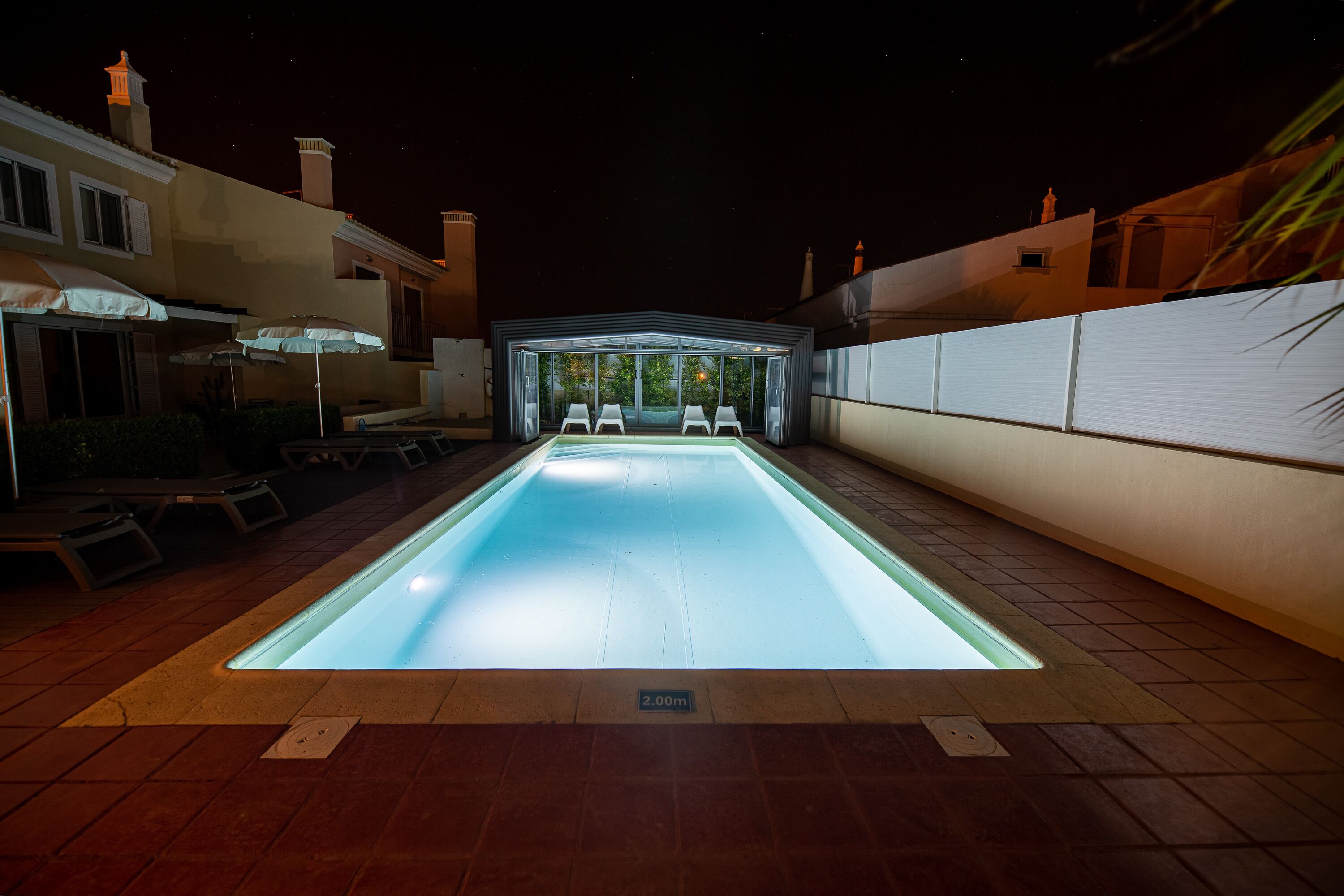 A rectangular swimming pool with a glass bottom, surrounded by a tiled deck and a covered patio area with chairs and umbrellas.
