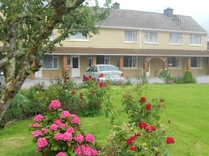 Una casa a due piani con un'auto nel vialetto, circondata da un giardino curato con fiori diversi e un prato.