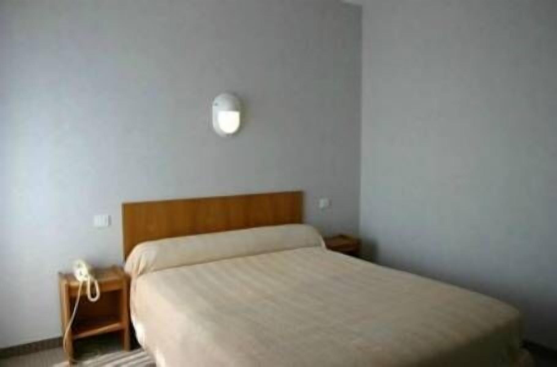 A simple, unadorned hotel room with a single bed, a bedside table, and a wall-mounted light fixture.