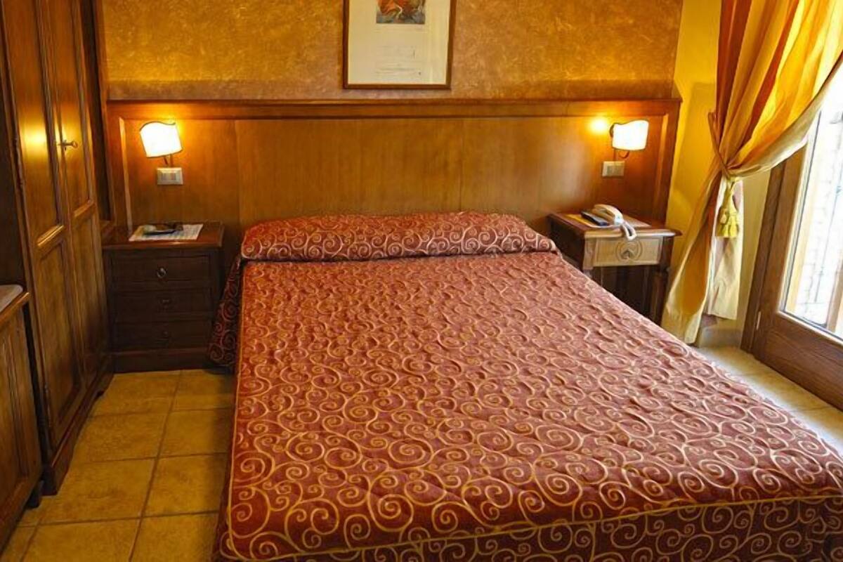 A hotel room with a bed, bedside tables, a nightstand, a desk, and a window with curtains.