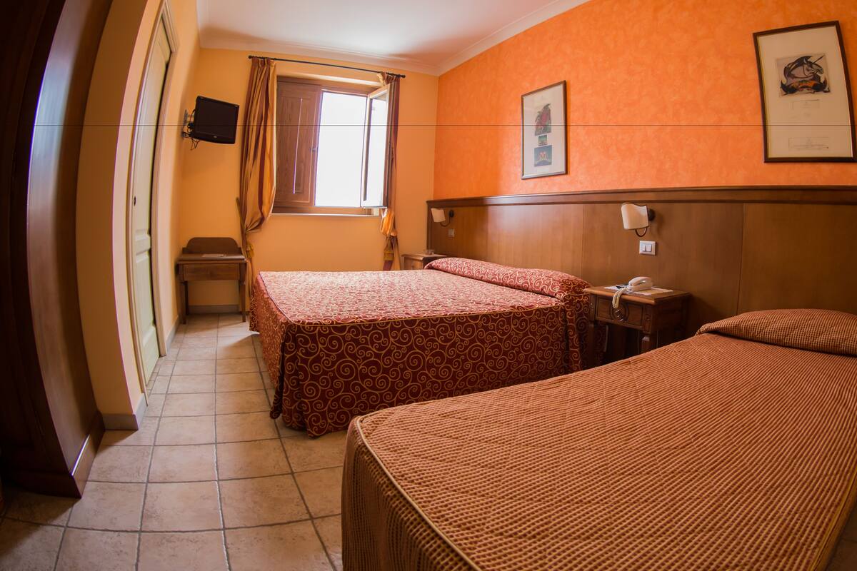 A hotel room with two single beds, a wooden headboard, a window with curtains, a wall-mounted TV, and a framed picture.