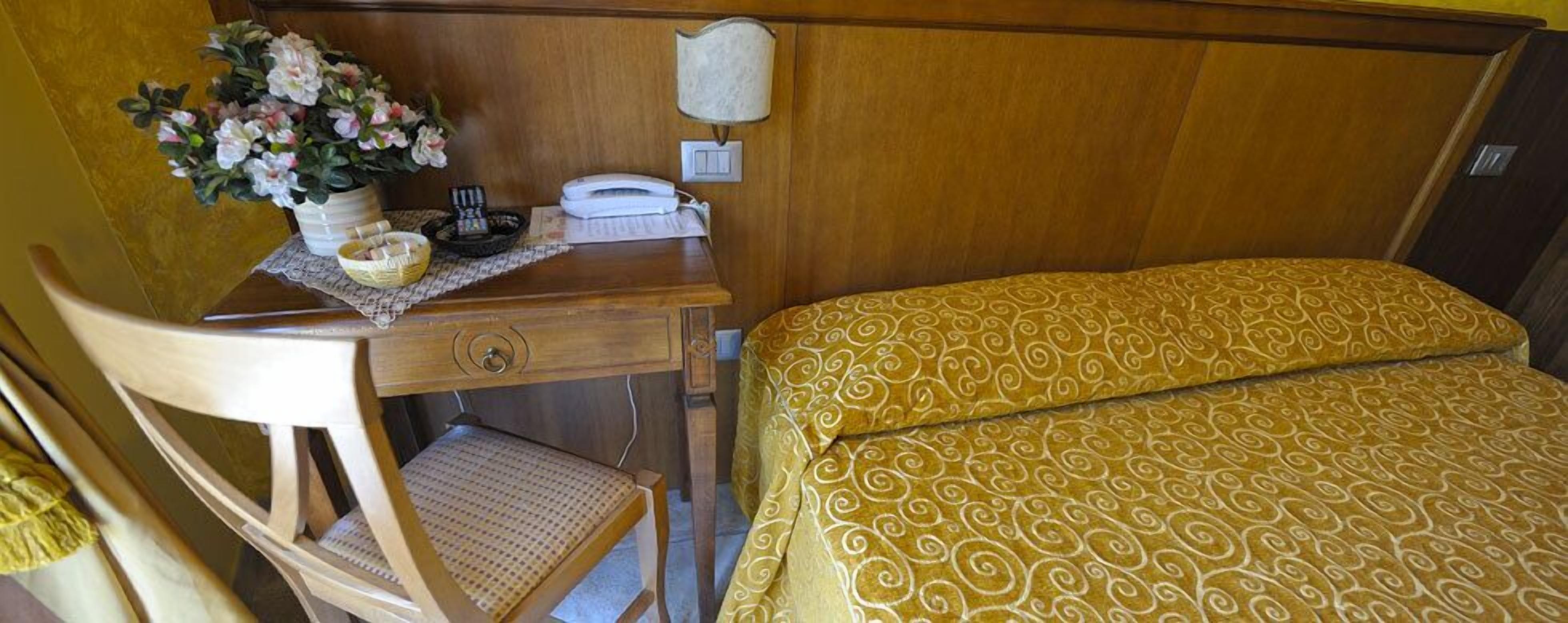 Habitación de hotel con escritorio de madera, teléfono, lámpara, cama con colcha estampada y silla con asiento de mimbre.