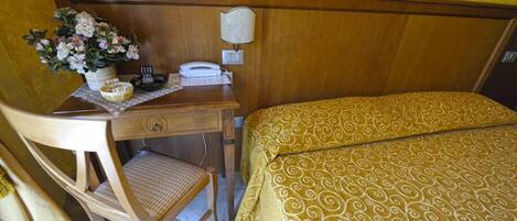 Hotelkamer met een houten bureau, telefoon, lamp, bed met een gestreept beddegoed en een stoel met een gevlochten zitting.