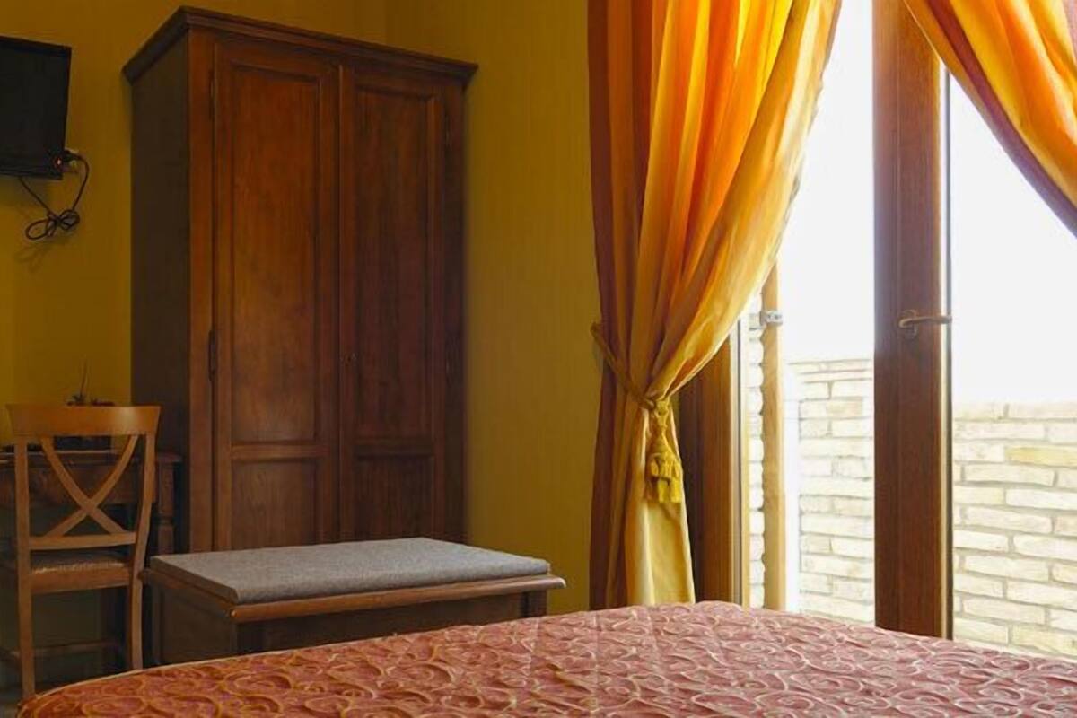 A room with a bed, a wooden wardrobe, a small table with a chair, a television mounted on the wall, and a window with curtains.