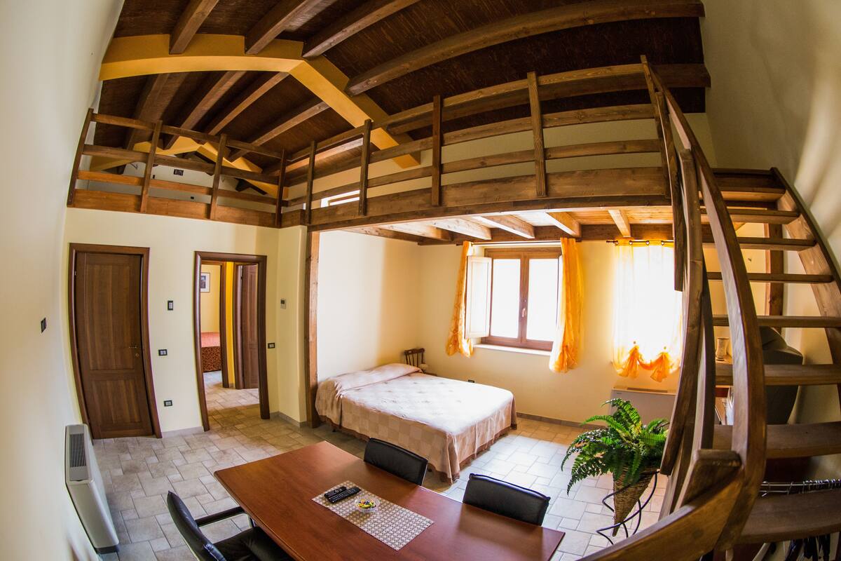 A room with a wooden ceiling, a bed, a table with chairs, and a staircase.