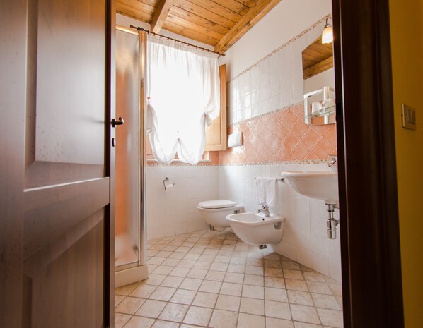 Un baño con inodoro, bidé y lavamanos, suelo de baldosas, techo de madera y una ventana con cortina.