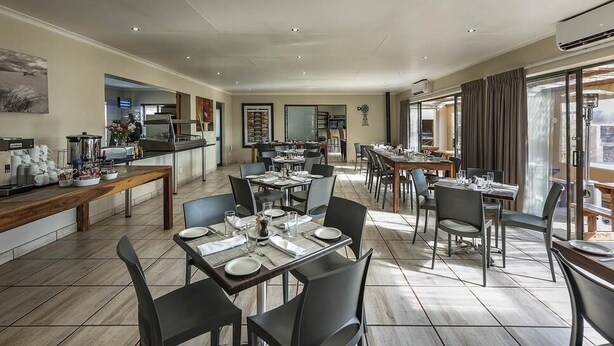 A spacious dining area with multiple tables and chairs, a kitchen in the background, and large windows providing natural light. - Kathu Inn (Kathu)