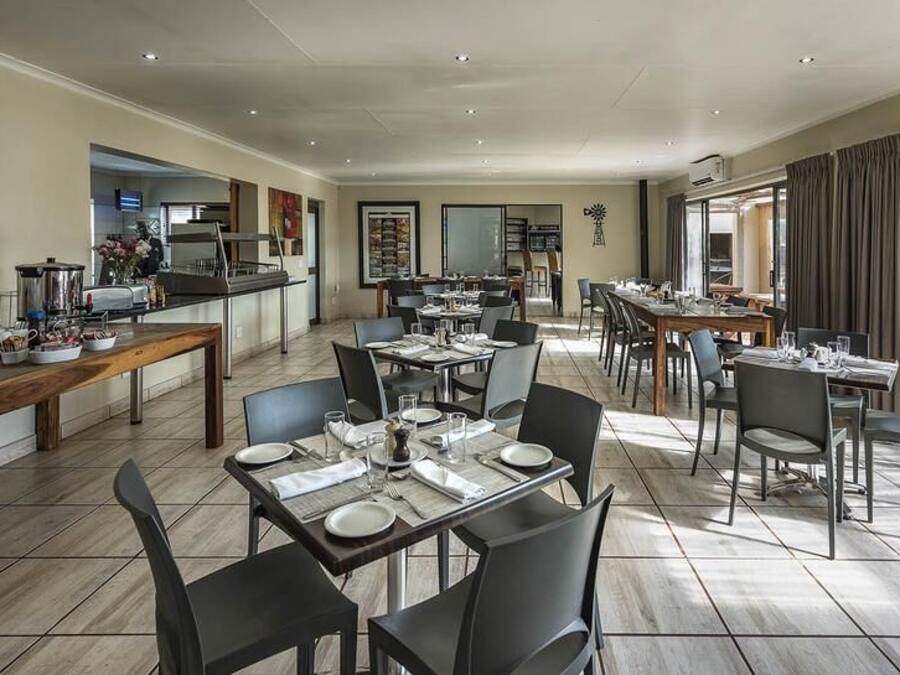 Uma ampla área de refeições com várias mesas e cadeiras, uma cozinha ao fundo e grandes janelas que proporcionam luz natural.