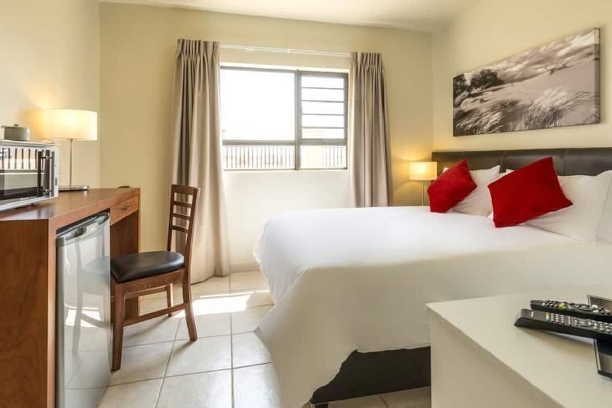A hotel room with a bed, a desk with a microwave, a chair, a TV, and a window with curtains.