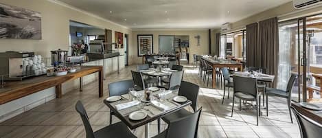 A spacious dining area with multiple tables and chairs, a kitchen in the background, and large windows allowing natural light.