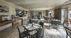 A spacious dining area with multiple tables and chairs, a kitchen in the background, and large windows allowing natural light. - Kathu Inn (Kathu)