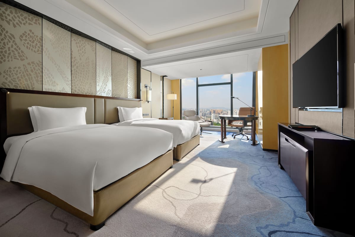 A hotel room with two beds, a desk, a chair, a TV, and a large window offering a city view.
