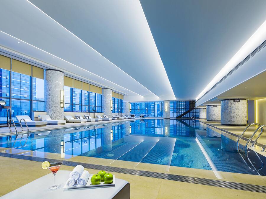 A modern indoor swimming pool with clear blue water, sun loungers, and a bar area with a drink and fruit.