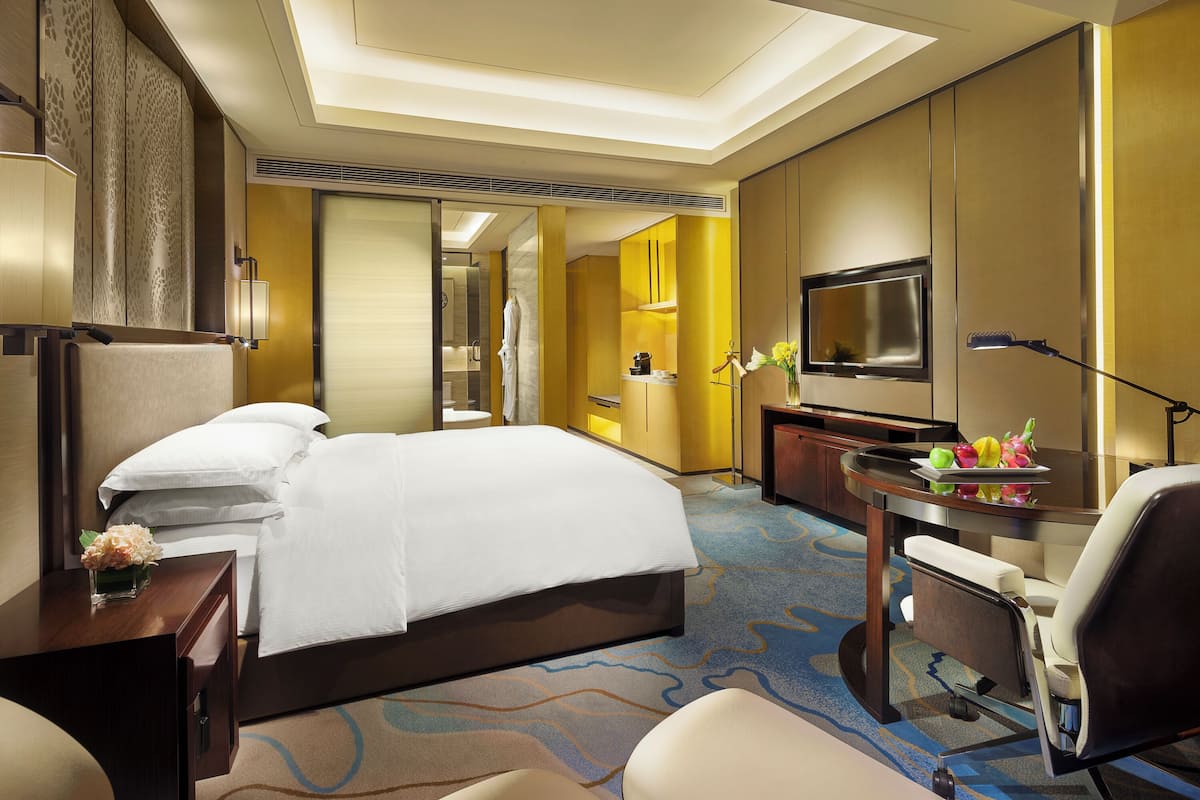 A hotel room with a large bed, a desk with a lamp, a TV, and a small table with fruit.