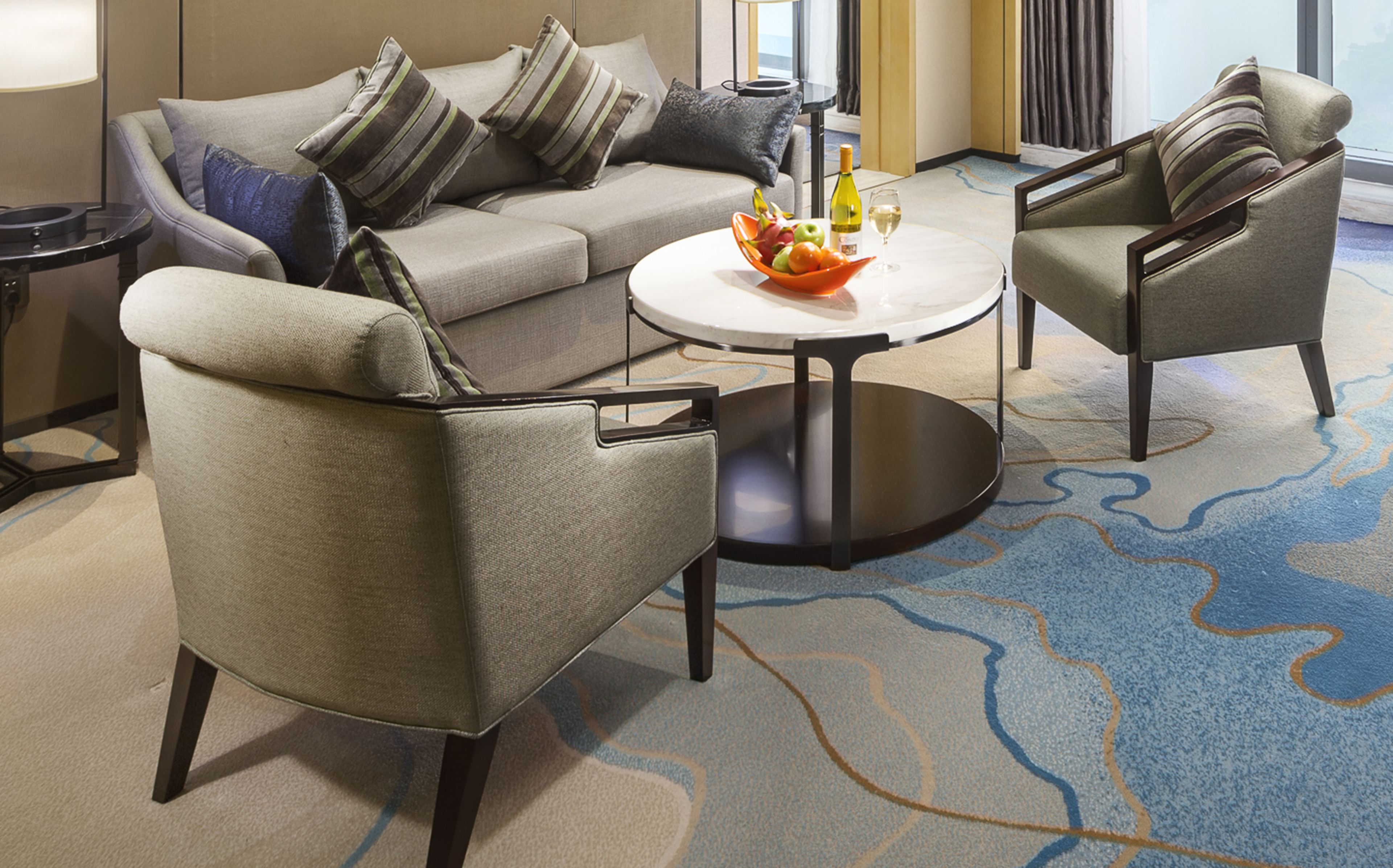 A modern hotel room with a grey sofa set, two armchairs, a round coffee table, and a fruit bowl.