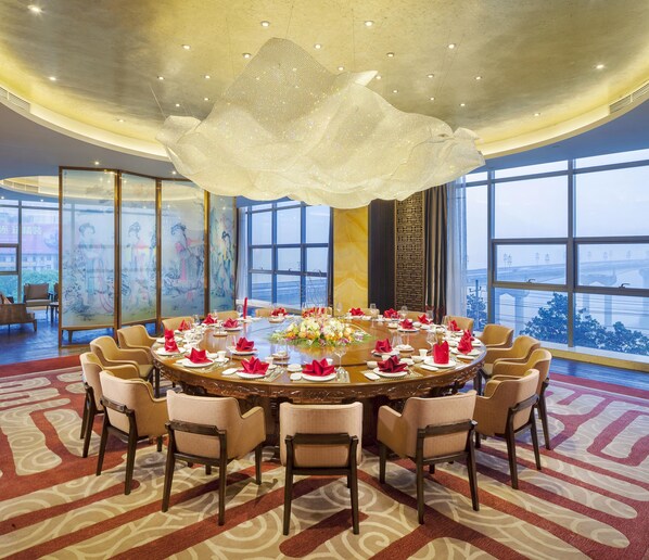 A round dining table set for a formal meal, surrounded by chairs, with a large chandelier overhead and a view of a bridge through large windows.