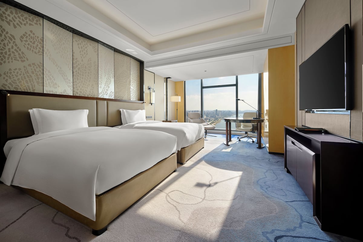 A hotel room with two beds, a desk, a TV, and a large window offering a city view.