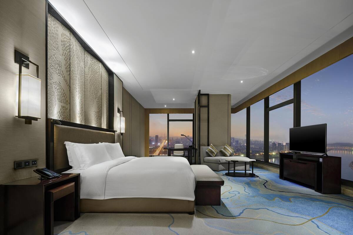A hotel room with a bed, a desk, a TV, a chair, and a large window with a city view.