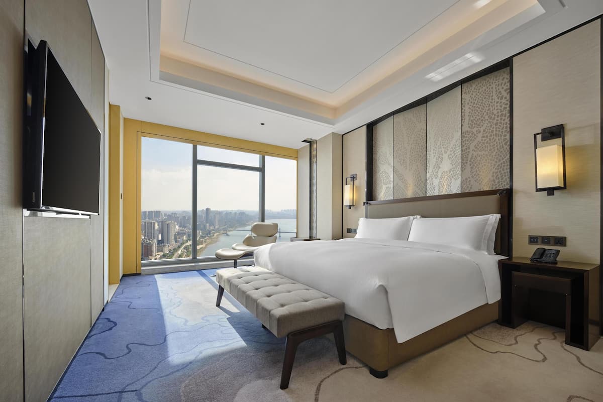 A modern hotel room with a large bed, a TV, a bench, a nightstand, and a city view.