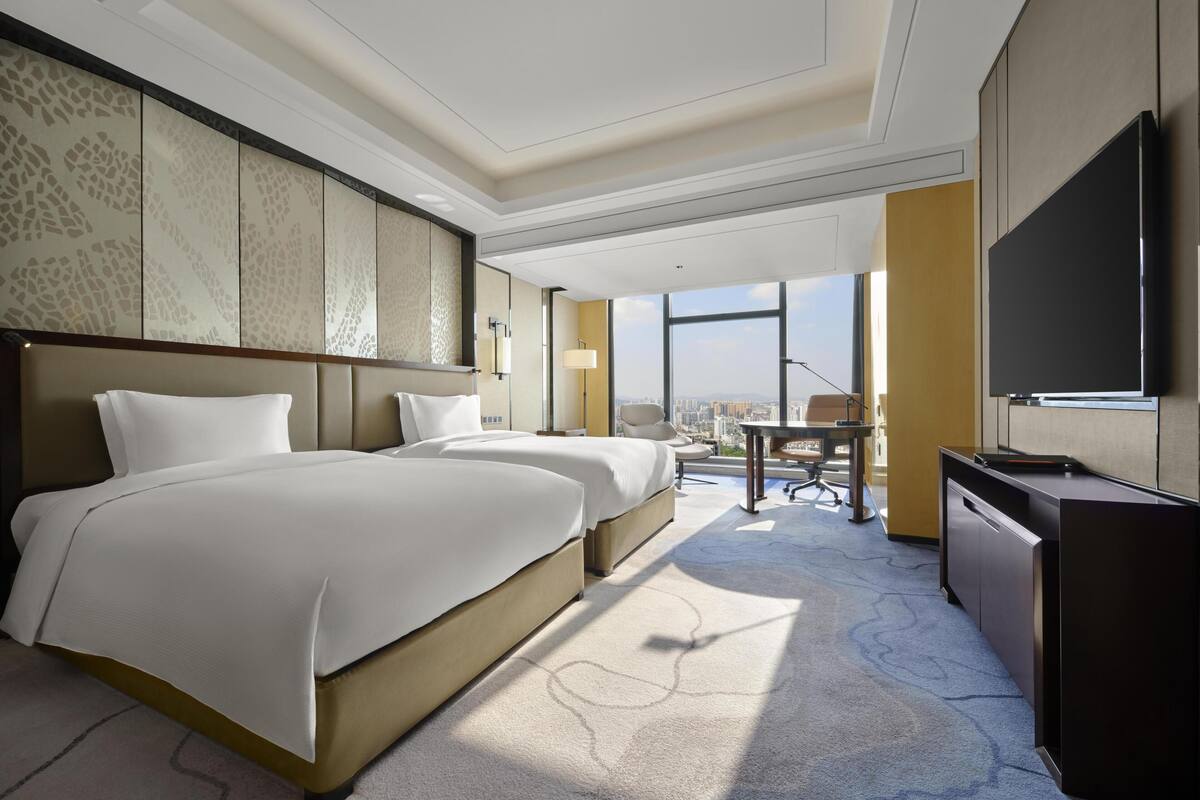 A hotel room with two beds, a desk, a chair, a TV, and large windows offering a city view.