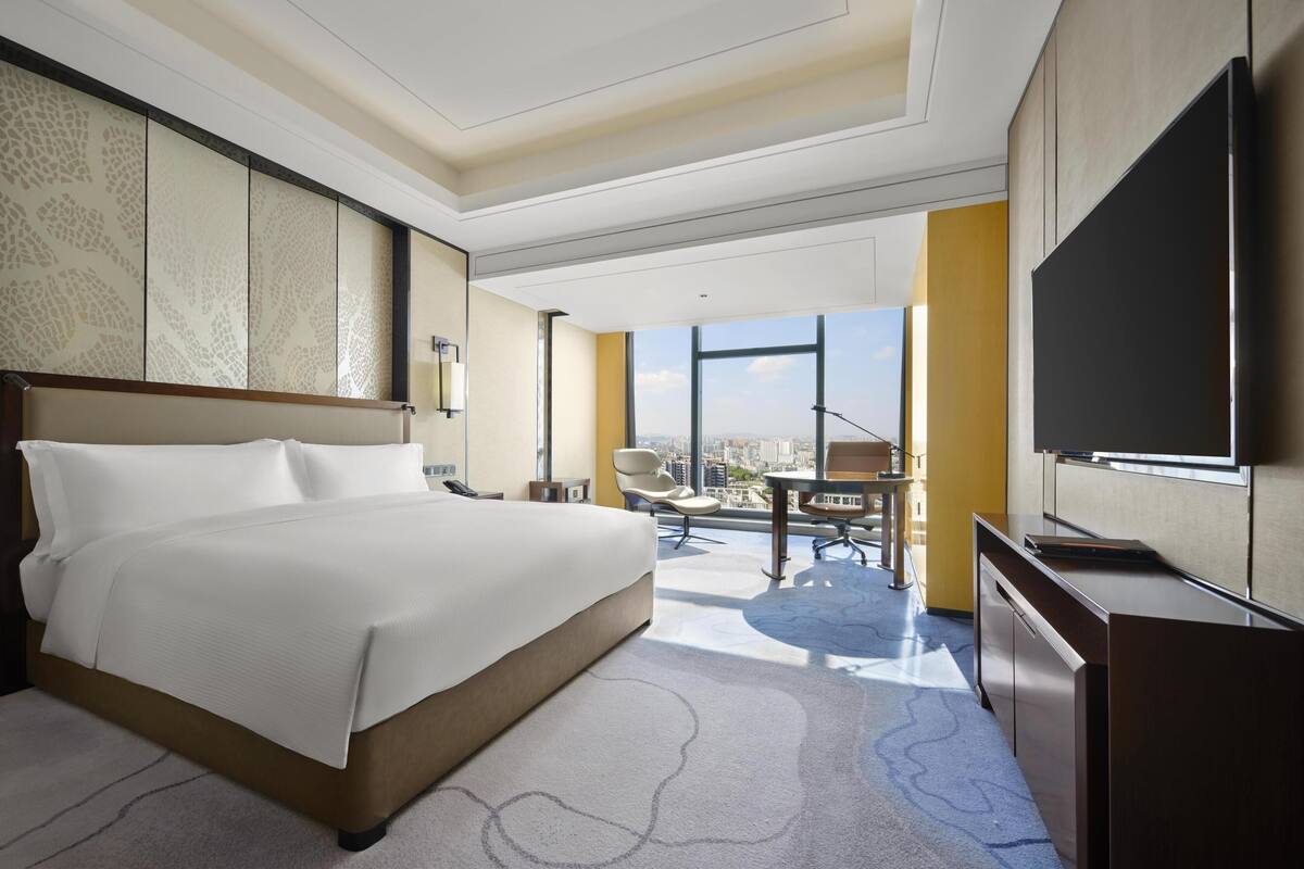 A hotel room with a large bed, a desk, a chair, a television, and a city view.