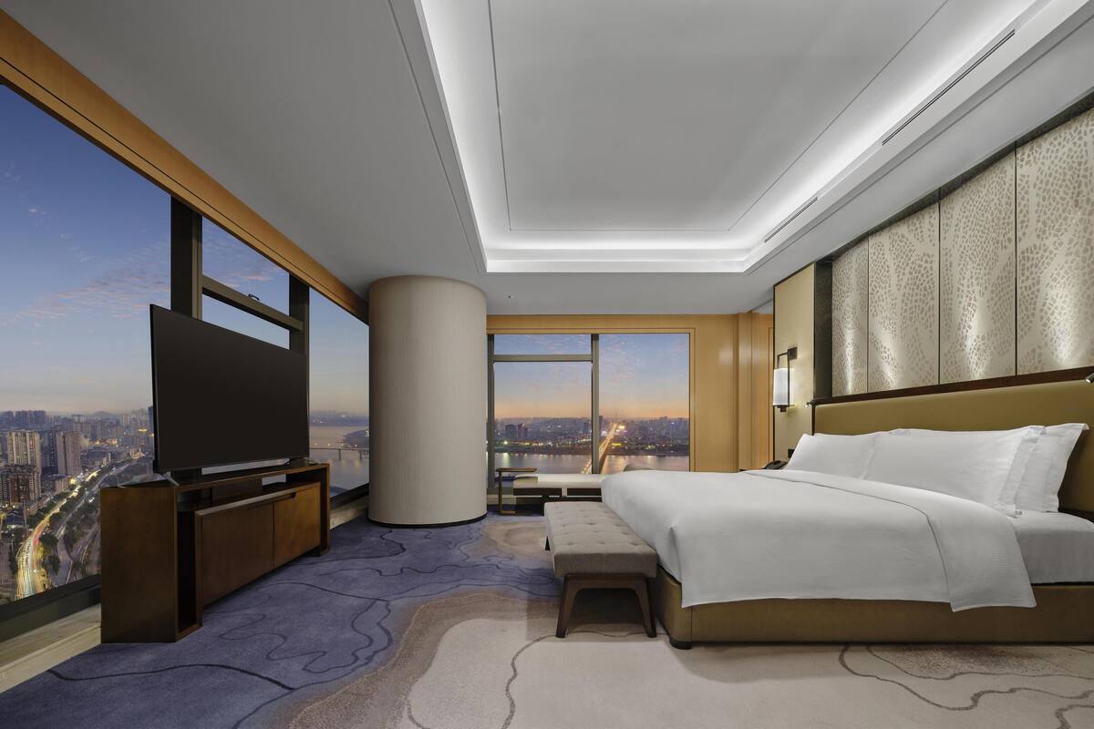 A modern hotel room with a large bed, a TV, a view of the cityscape, and a large window.